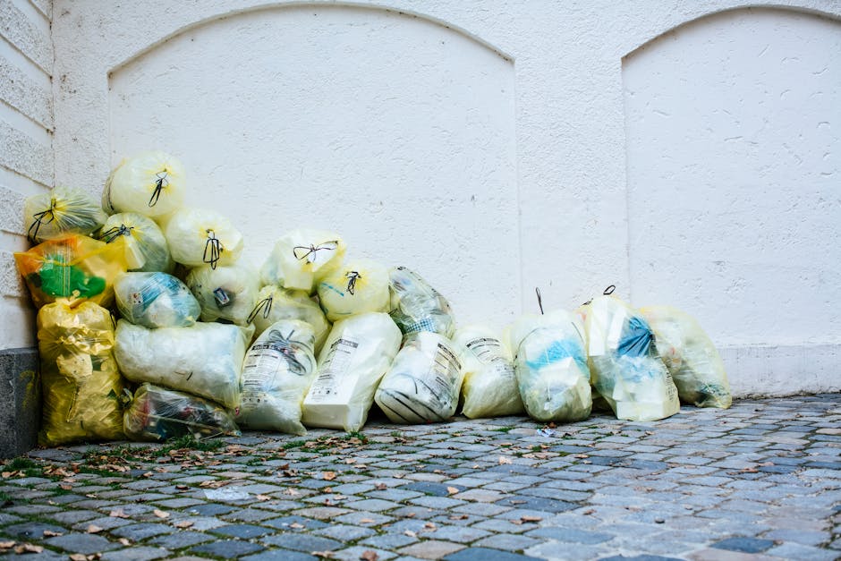 A collection of numerous plastic rubbish bags, mostly in yellow, white, and transparent tones, are stacked against a white exterior wall near a paved cobblestone surface. The bags appear to be filled with various household waste materials, with some showing labels or printed text on the plastic. The bags are positioned on the ground, with some leaning against the stone wall on the left side and others stacked informally in front of a plain white wall, suggesting an area designated for waste collection outside a building. The surface underneath consists of small, dark cobblestones that contrast with the light-colored bags and wall, and there are a few scattered leaves and debris on the ground. The scene is illuminated with natural light, giving a clear view of the textures and colors of the plastic bags and surrounding environment. This arrangement is indicative of private or independent rubbish disposal, as it resembles on-site waste storage awaiting collection, consistent with services offered by companies like rubbishremovalwestkensington.com.