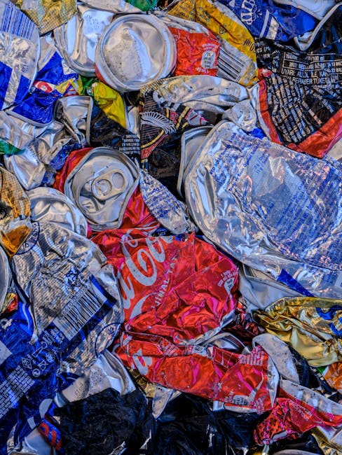 Close-up view of a pile of crushed aluminium cans and plastic soft drink bottles, primarily in silver, red, blue, black, and yellow colors. The aluminium cans feature various brand logos and text, with some showing recognizable designs like the Coca-Cola logo in red. The plastic bottles have textured surfaces with ridges and are slightly crumpled, with some caps visible attached to the bottles. The cans and bottles rest haphazardly on a surface that appears to be part of a waste container or recycling collection point, possibly outdoors given the natural diffuse lighting. The cluttered arrangement underscores the importance of proper waste disposal and highlights the type of irregular rubbish collection that private services like rubbishremovalwestkensington.com often handle, offering alternative waste management solutions for households and businesses. The image exemplifies common recyclable waste materials that require appropriate separation and disposal according to local regulations or through private rubbish removal services, supporting environmentally responsible waste management practices.