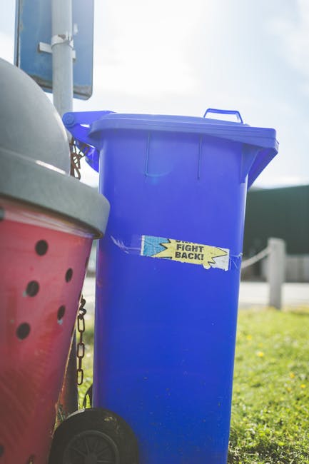 A white paper cup with printed text and green accents lies fallen on a paved ground outside, partially tilted with its open top facing up. The cup is situated on weathered, dark-colored bricks, which appear slightly uneven and have some dirt and debris scattered around. In the background, there is a section of a metallic drain cover with parallel slats, positioned near the cup, and a blurred outdoor environment with muted green and brown tones. The scene suggests a typical outdoor area where rubbish might be casually discarded, and it relates to private waste disposal or rubbish removal services, such as those offered by rubbishremovalwestkensington.com, emphasizing the importance of proper waste handling to avoid fines and adhere to local rubbish disposal rules.