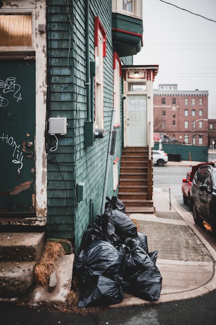 A group of black garbage bags filled with waste are piled at the base of a steep wooden staircase attached to a building with teal, shingle-style exterior siding. The bags rest on a small concrete landing adjacent to a sidewalk, near a weathered door with graffiti on its surface and a small set of concrete steps leading up to a dark red door. A white utility box and a black cable are mounted on the building's wall, and a small patch of dried grass is visible near the bottom left corner. In the background, there are parked cars and multi-storey brick buildings set against an overcast sky, with power lines overhead. The scene appears to be an urban residential area where private waste collection or rubbish removal services, like those offered by Rubbish Removal West Kensington, might be used to handle non-standard waste disposal outside of local authority services.