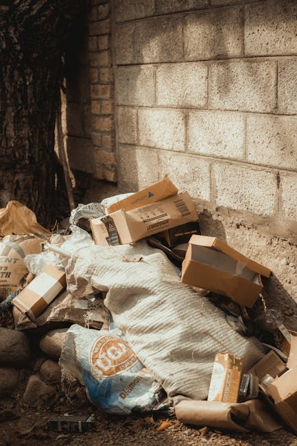 A black, wheeled rubbish trolleys with a large fabric bag closure, positioned in the centre of a narrow urban alleyway, surrounded by piles of mixed waste materials including cardboard boxes, plastic bags, and discarded packaging at the back. The alley is flanked by graffiti-covered walls on the right and weathered, brick and metal surfaces on the left, with some exposed pipelines and electrical conduits visible. In the background, an assortment of damaged cardboard boxes, paper debris, and plastic waste is stacked irregularly against a makeshift wall, suggesting accumulation of waste typical of private refuse collection or on-site clearance. The ground is dark, uneven, and stained, indicative of frequent use for rubbish disposal. The scene appears to be outdoors, lit by natural daylight, with bare tree branches overhead and surrounding buildings that imply an inner-city setting where waste might be temporarily stored before disposal, aligning with practices beyond municipal rubbish removal services, as addressed in the context of private waste handling and rubbish removal services by rubbishremovalwestkensington.com.