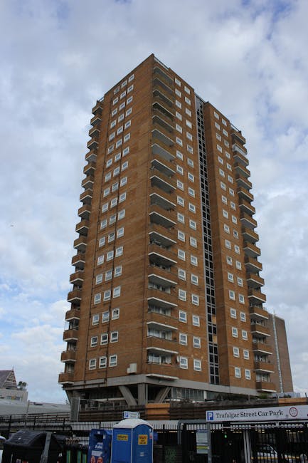 A tall, multi-storey residential building made of reddish-brown brick, featuring numerous windows and small balconies on each floor. The structure has a modern, utilitarian design with a central vertical section of larger windows and a dark vertical strip running along the façade. The building is situated on a paved urban street, with a fenced parking area at the base that includes a sign for 'Trafalgar Street Car Park' and a row of portable toilets nearby. The sky overhead is partly cloudy, creating diffused natural lighting that highlights the building's textures and architectural details. This scene captures an environment where private waste management or rubbish removal services, such as those provided by companies like rubbishremovalwestkensington.com, may carry out on-site clearance of rubbish from residential flats. The setting illustrates the typical context of high-rise waste disposal activity in an urban West Kensington environment, with no visible waste or debris in the immediate vicinity.