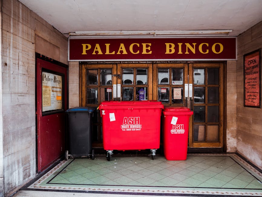 Inside the entrance of Palace Bingo, a public entertainment venue, there are three waste containers positioned on a tiled floor in front of a wooden-framed glass doorway. The largest container is a red wheelie bin with a white and red label that reads 'ASH Waste Services,' designed for general rubbish disposal, with a slightly textured plastic surface. To its right, a smaller red bin, also labeled for waste collection, stands on four wheels, with a smooth surface and a similar bright red colour. On the left, a black wheelie bin with a matte finish, used for different waste streams, is visible. Behind these containers, the wooden-glass door setup features multiple small square glass panes, some of which have notices or signs attached. Above the door, a large sign reads 'PALACE BINGO' in prominent yellow lettering on a maroon background. The surrounding environment includes walls with aged, beige-colored tiles and a red-framed notice board on the right side of the entrance, suggesting an urban setting. The lighting appears to be soft, ambient, with natural or indoor light giving a clear view of the waste containers and immediate surroundings. The image, relevant to private waste handling, implies the venue's open policy, with these waste bins likely used for on-site rubbish collection separate from municipal services, supporting alternative rubbish removal approaches.