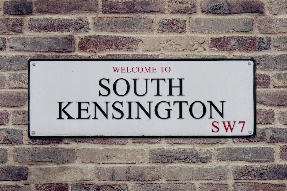 A rectangular metal street sign mounted on a brick wall, displaying the text 'Welcome to South Kensington SW7' with 'South Kensington' in large, bold black letters, and 'Welcome to' and 'SW7' in smaller red font. The sign has a white background with rounded corners and four visible screws securing it to the wall. The brick wall behind is composed of reddish-brown bricks with a textured surface and visible mortar joints, providing a classic urban setting. The lighting is natural, highlighting the contrast between the sign's clean, smooth surface and the rougher texture of the bricks. This scene is typical of signage used in urban residential or commercial areas, subtly supporting the context of local place identification, which in turn connects to services such as private rubbish removal for properties in South Kensington. The image emphasizes clarity, outdoor environment, and the integration of functional signage within a historic brick façade, aligning with themes of on-site waste management and alternative disposal options for urban properties.