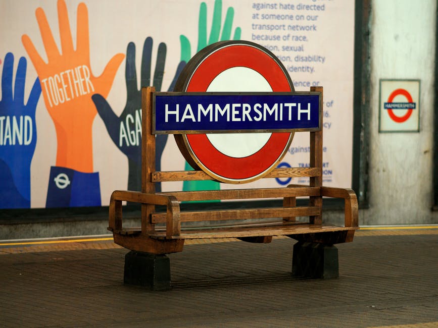 A wooden bench with a backrest and armrests, situated on a dark paving surface, features a sign with a blue background and white lettering reading 'HAMMERSMITH' in front of a circular London Underground entrance sign with a red border, white center, and black frame. In the background, a colorful mural with stylized hand drawings in orange, blue, and green, accompanied by text, is visible on a wall. There is also a smaller round sign with a red border, white center, and blue text reading 'Hammersmith,' mounted on a white wall to the right. The scene appears to be inside a London Underground station, illustrating a setting related to urban transport and local area identification, with an emphasis on clean, well-maintained environment and visual cues for commuters.