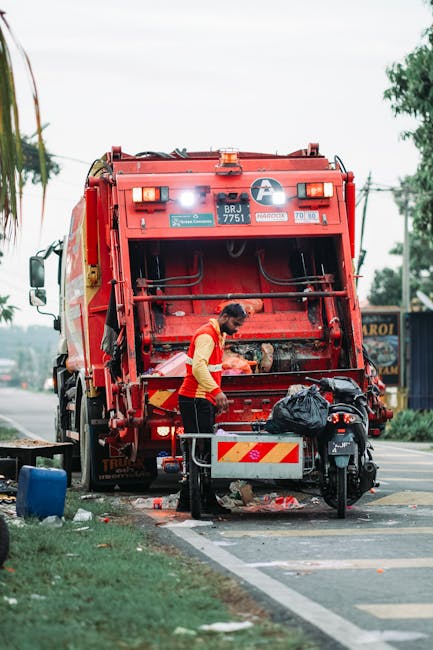 A waste collection worker wearing a yellow high-visibility vest and black trousers is operating a red rubbish truck positioned on a street, with the rear hatch open. The worker appears to be managing waste materials, with a black plastic bag placed on a nearby platform attached to a refuse collection bike. The truck is equipped with orange warning lights, and the vehicle’s body features reflective safety markings. The scene is set outdoors during daytime, with an overcast sky providing diffused lighting. In the background, there are trees and some distant buildings, indicating an urban or suburban environment. The street has a visible curb and part of a designated parking or loading lane, with some scattered rubbish and a few small objects nearby, emphasizing the context of street cleaning or rubbish removal services. The overall scene captures a routine operation of private waste disposal, aligned with independent rubbish removal practices that may be preferred as an alternative to local authority collection. The presence of the company name or logo is not visible, but the image clearly illustrates rubbish collection activity typical of West Kensington areas seeking professional waste management. This detailed description supports accessibility and SEO relevance for content about rubbish collection services. 