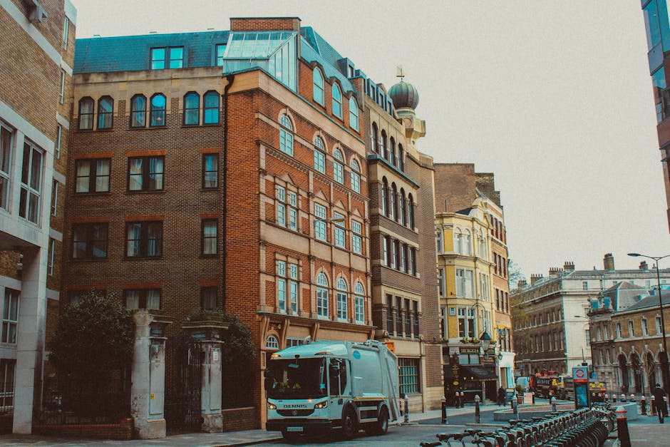 A street scene in an urban area featuring multi-story buildings with diverse architectural styles, including brick facades, decorative window frames, and a rounded tower with a domed roof topped by a weather vane. In the foreground, a white waste collection vehicle is parked along the curb, positioned between a stone gate and a row of parked bicycles secured to a metal bike rack. The vehicle's side panel and front are visible, with some orange safety equipment on top. To the right, there are additional buildings with large windows and detailed masonry, while the street is lined with bollards, lampposts, and traffic signs, indicating a busy city environment with active pedestrian and vehicle movement. The lighting appears natural, suggesting a daytime setting with slightly overcast skies. This scene reflects an aspect of private waste collection, typical of independent rubbish removal services in central urban areas, such as West Kensington, aligning with practices of on-site clearance and alternative waste handling methods. The focus on the waste collection vehicle highlights the context of rubbish removal services working in city streets, with buildings serving as the backdrop for everyday urban waste management operations.