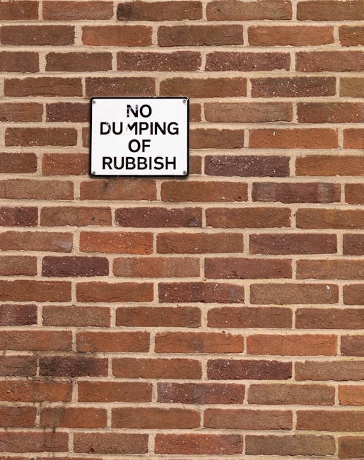 A rectangular white and black metal sign mounted on a reddish-brown brick wall displaying the message 'NO DUMPING OF RUBBISH' in bold black text. The bricks are arranged in a horizontal pattern, with a mix of lighter and darker shades, and the surface of the wall appears textured. There are no other objects or environmental features visible in the image, indicating the sign's purpose to deter illegal rubbish disposal in this area, consistent with regulations on private waste handling or onsite clearance practices. The image emphasizes the importance of proper rubbish management and the role of signage in supporting waste removal services such as those offered by companies in the vicinity of West Kensington. The natural lighting highlights the brick textures and the clarity of the sign.