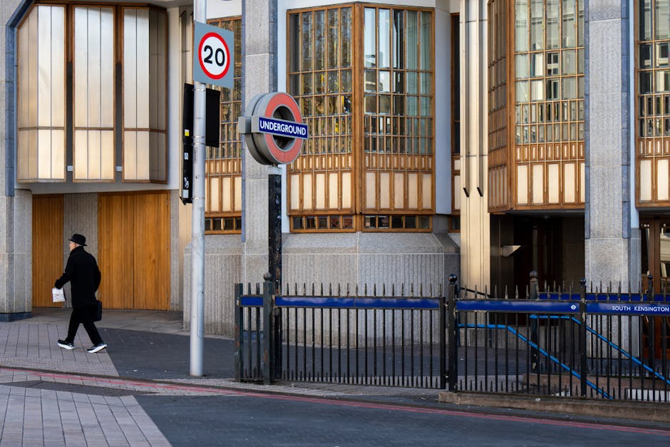 Exterior view of a modern building with large wooden-framed windows and concrete panels on the facade, situated along a sidewalk at South Kensington underground station. A black metal railing runs parallel to the curb, partially enclosing an underground entrance surrounded by concrete walls. In the foreground, a person dressed in dark clothing and a hat walks along the pavement, carrying a white paper bag. There are two road signs visible: a red and white circular speed limit sign indicating '20', and a vertical rectangular sign with the classic underground symbol and the word 'UNDERGROUND' in white on a blue background. The scene is lit with natural daylight, highlighting the textures of the wooden window frames, the concrete surfaces, and the pavement pattern, which consists of grey and pinkish bricks. The environment appears tidy and well-maintained, with elements relevant to urban transport hubs and private waste handling reflecting the area's infrastructure.